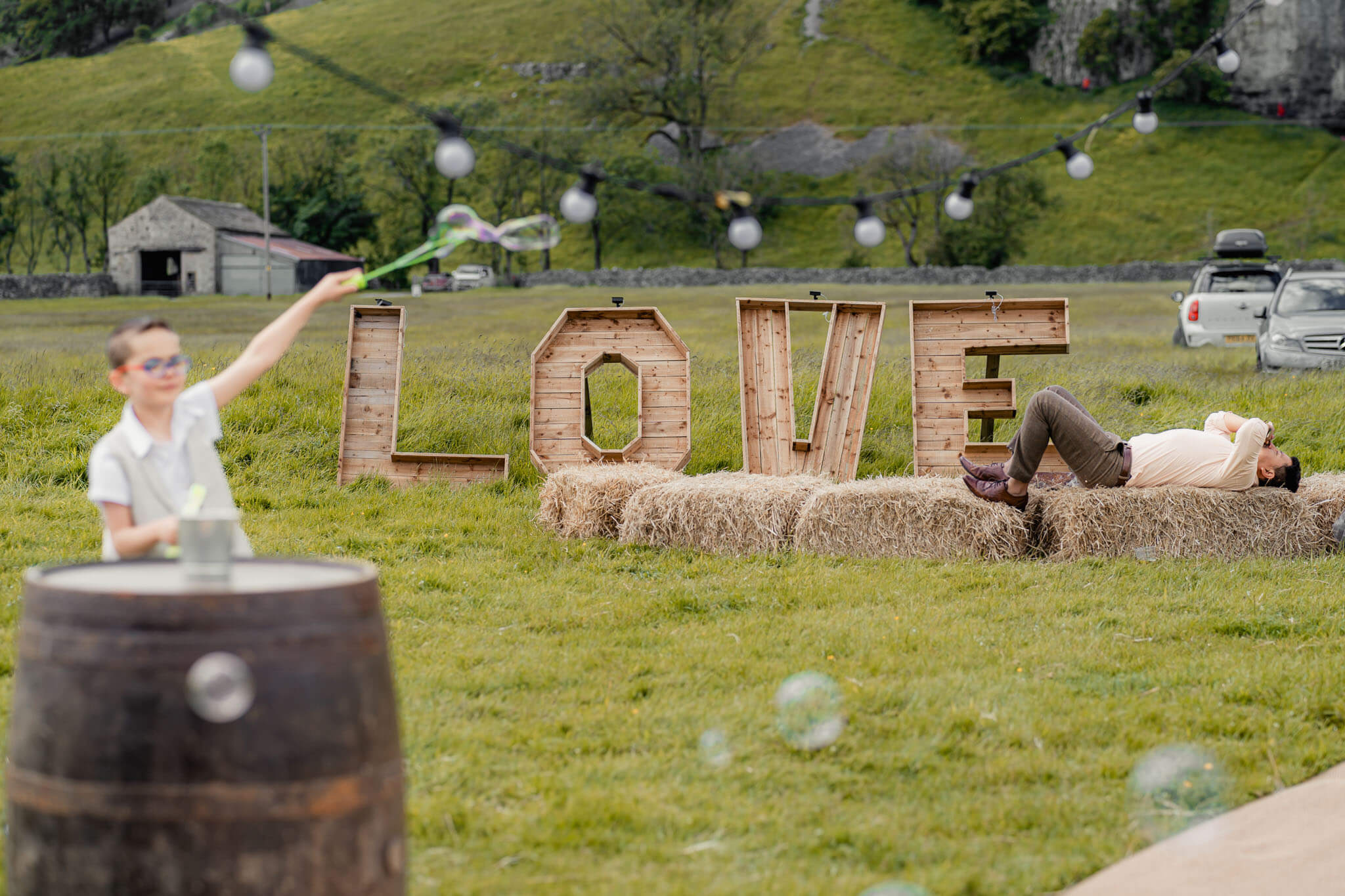 A fun and relaxed outdoor wedding setting featuring a wooden "LOVE" sign on hay bales, a playful child blowing bubbles, and a guest lying back enjoying the moment.