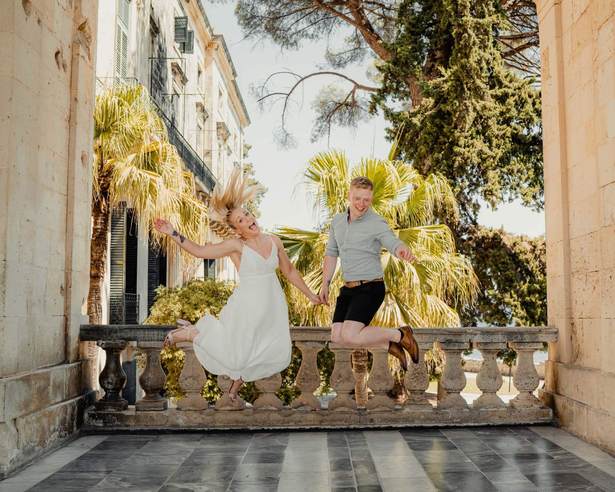 A joyful couple jumping in excitement, holding hands in a beautiful outdoor setting with palm trees and historic architecture in the background.