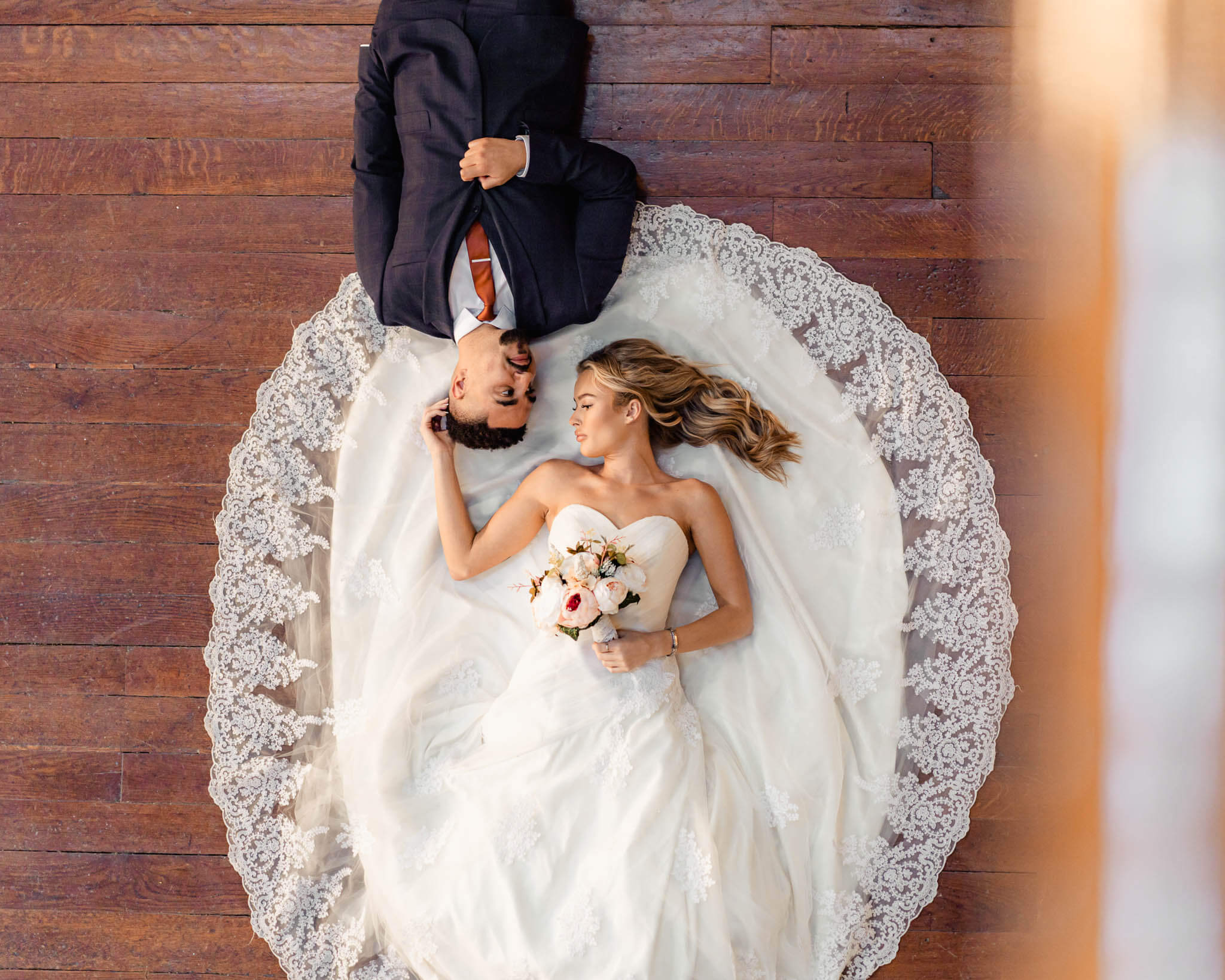 A bride and groom lying on a wooden floor, with the bride’s lace gown elegantly fanned out around them, sharing a peaceful and intimate moment.