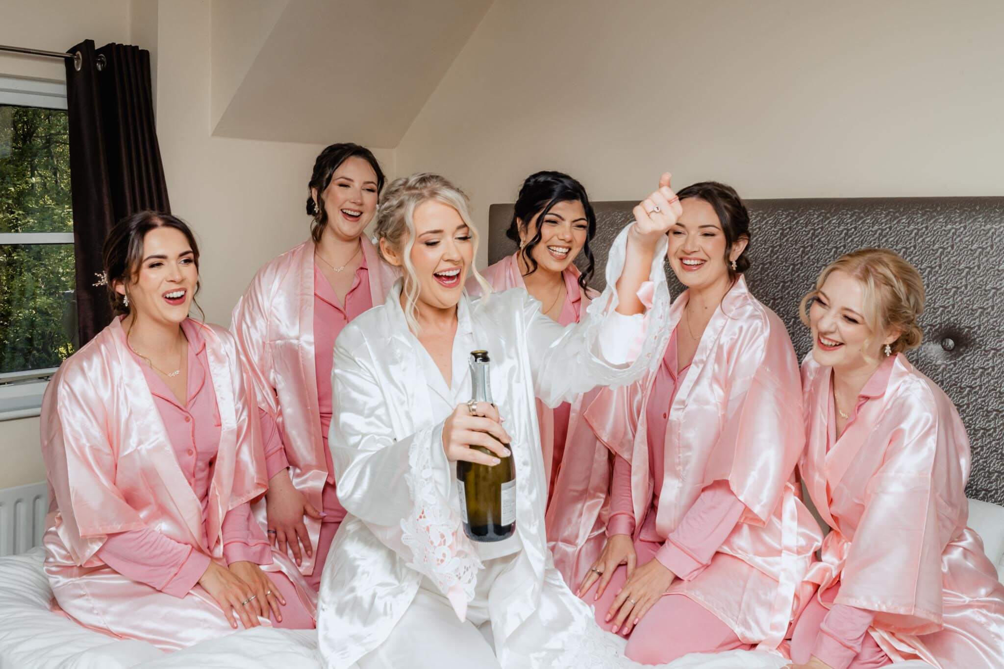 Bride popping champagne surrounded by laughing bridesmaids in pink robes.