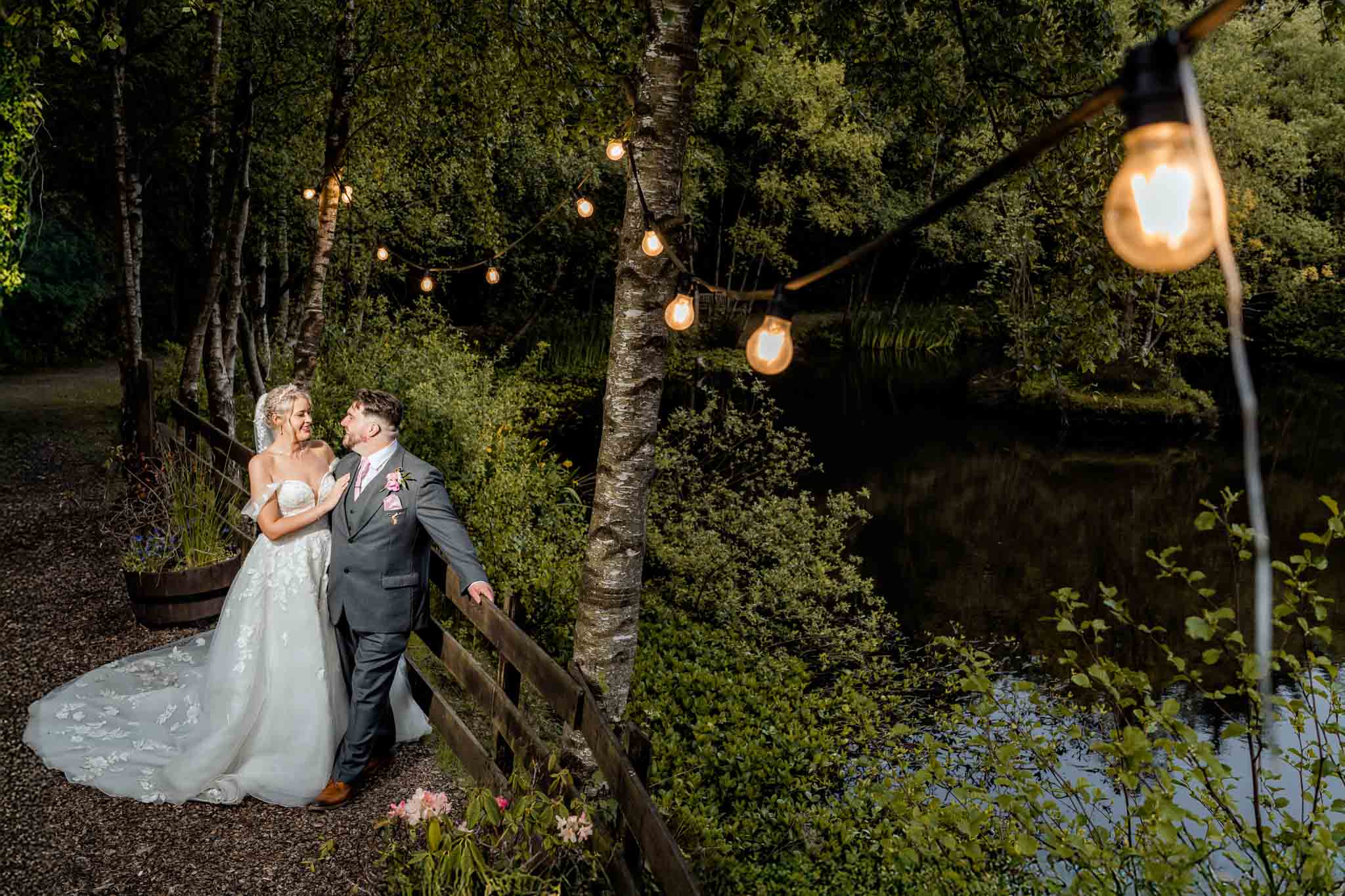 Bride and groom smiling under fairy lights by the lake.