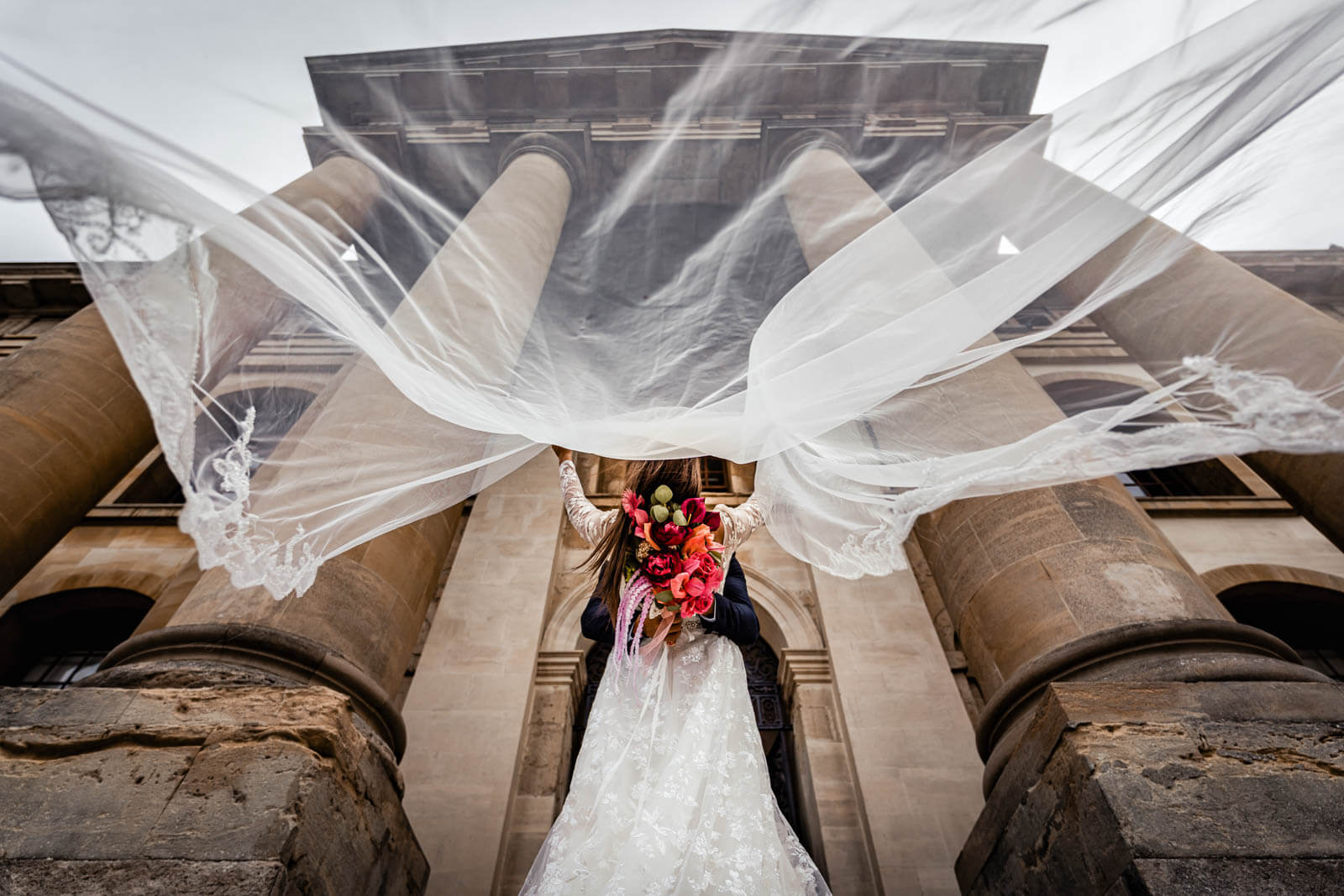 Bride and groom embrace beneath flowing veil in front of grand stone columns - wedding photography styles in Yorkshire.