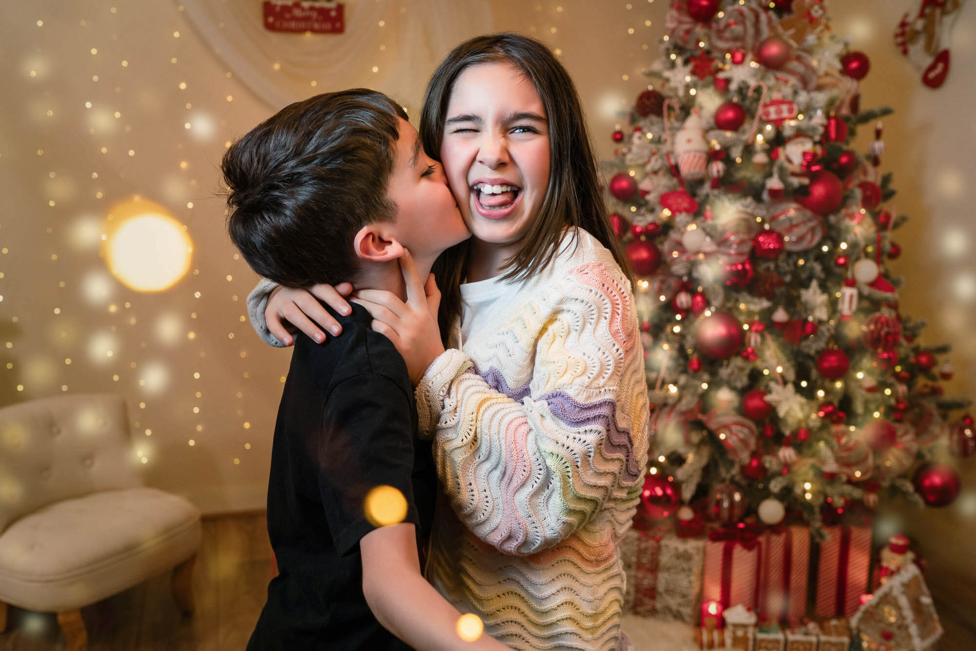 Brother kissing sister during behind-the-scenes Christmas session with red tree décor in Yorkshire.