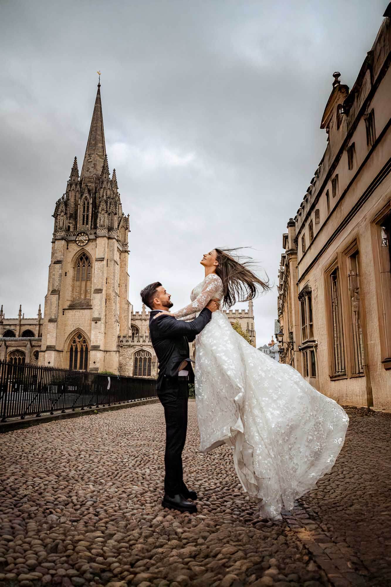 Groom lifting his bride in front of Oxford architecture in an elegant editorial wedding portrait.