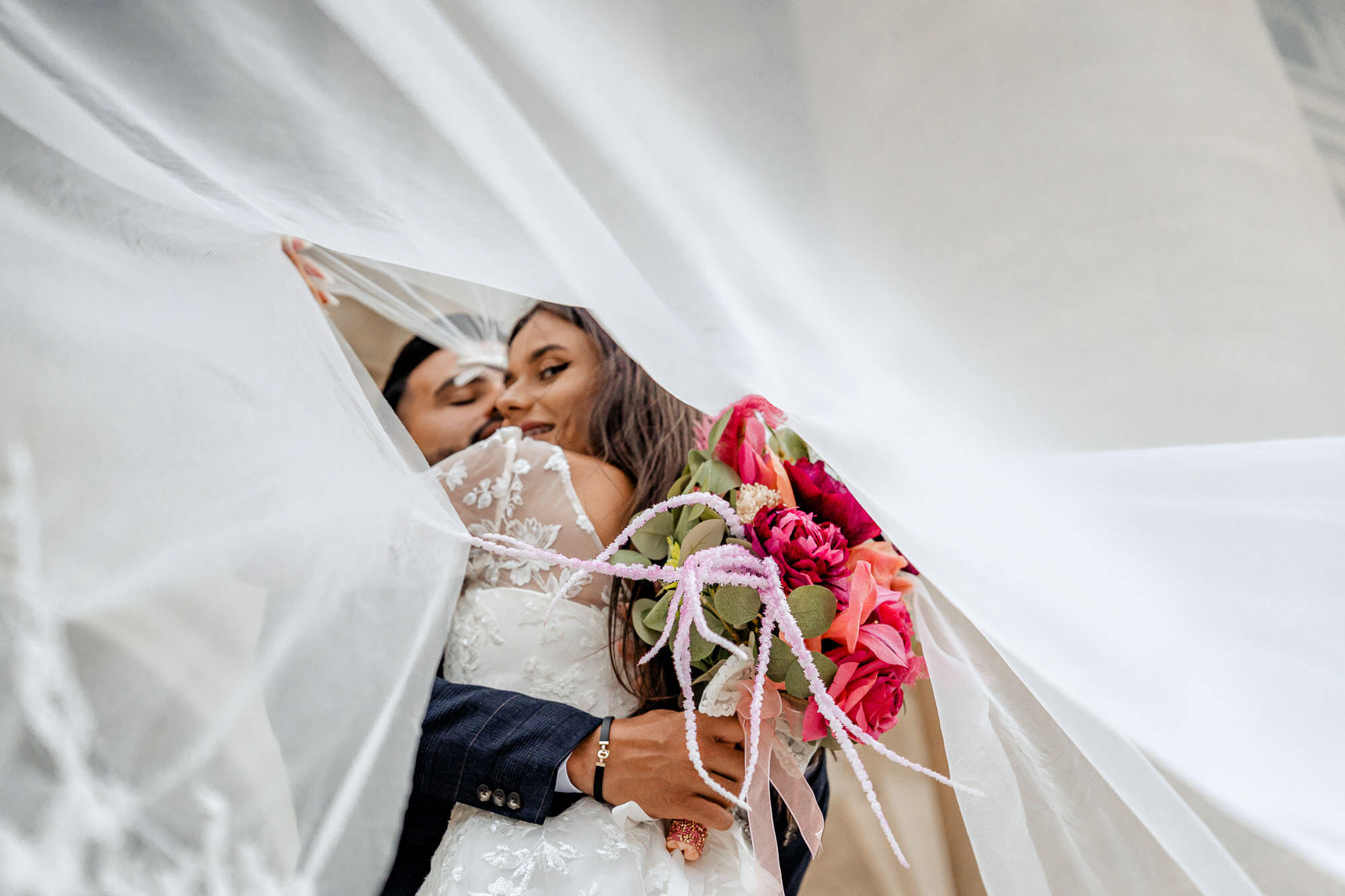 Creative close-up of a couple embracing under the veil with a vibrant pink wedding bouquet.