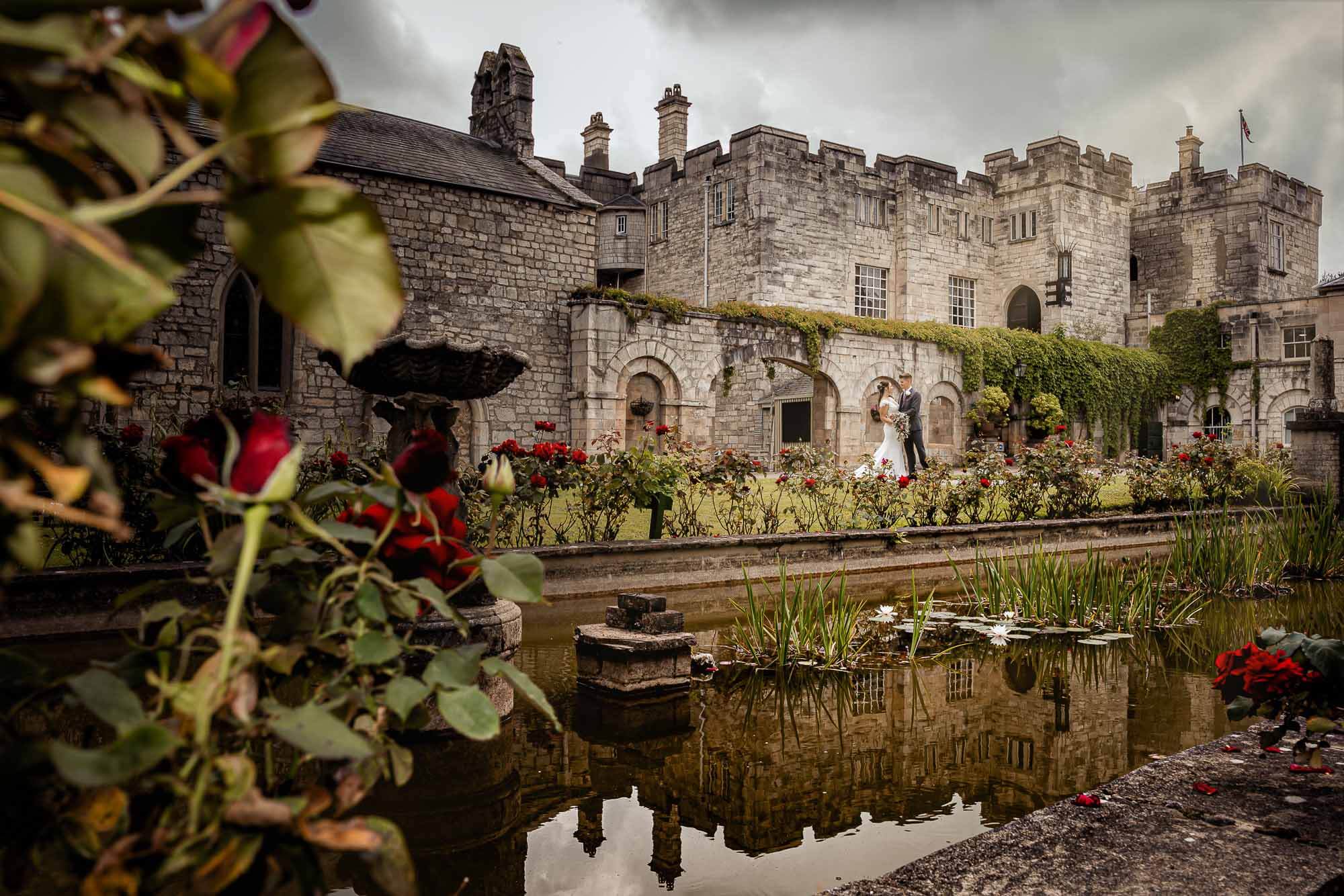 Bride and groom walking through historic castle gardens reflected in a still pond.