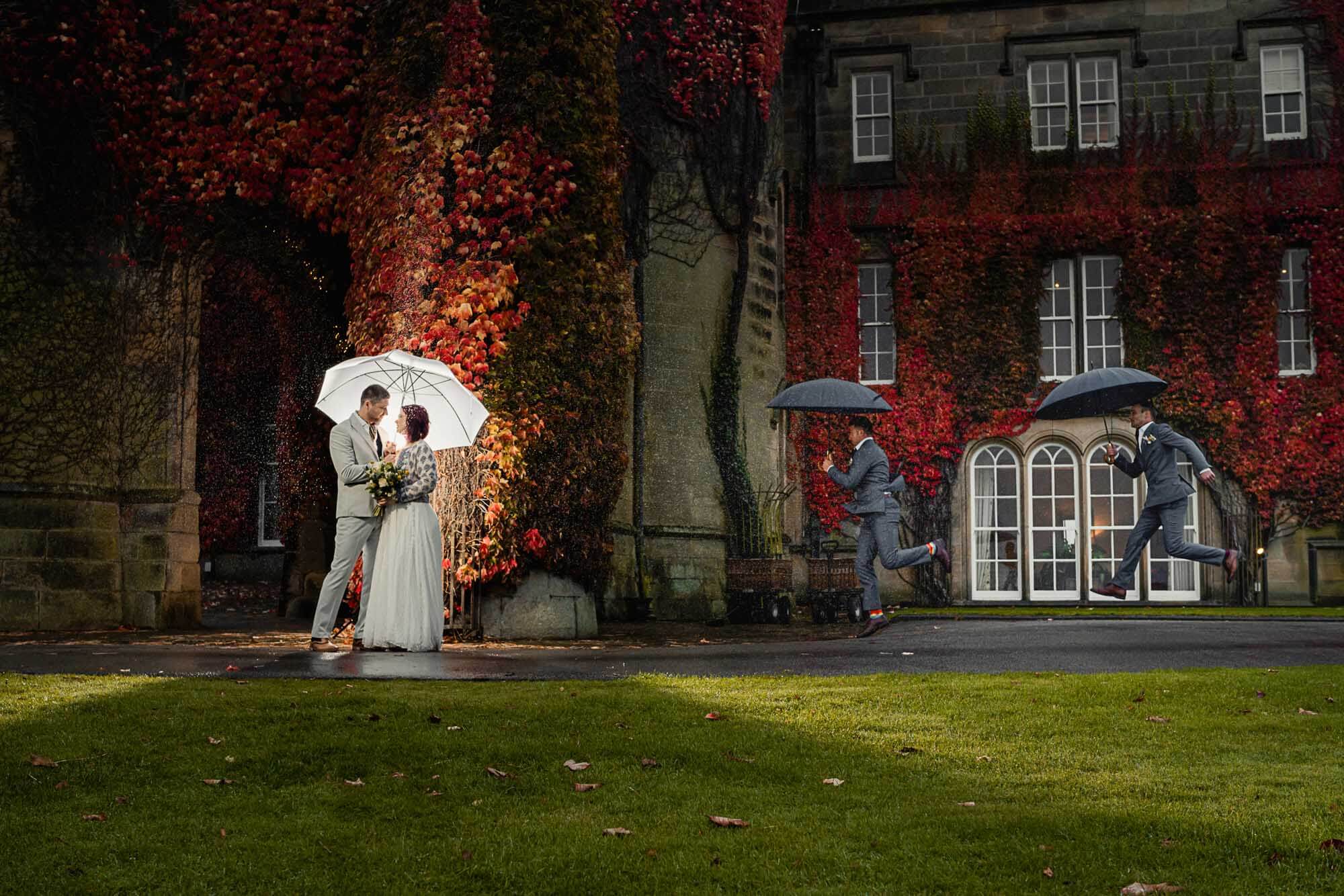 Cinematic elopement wedding in Yorkshire as the couple stand under an umbrella in the rain, with playful motion and autumn ivy covering the historic building.
