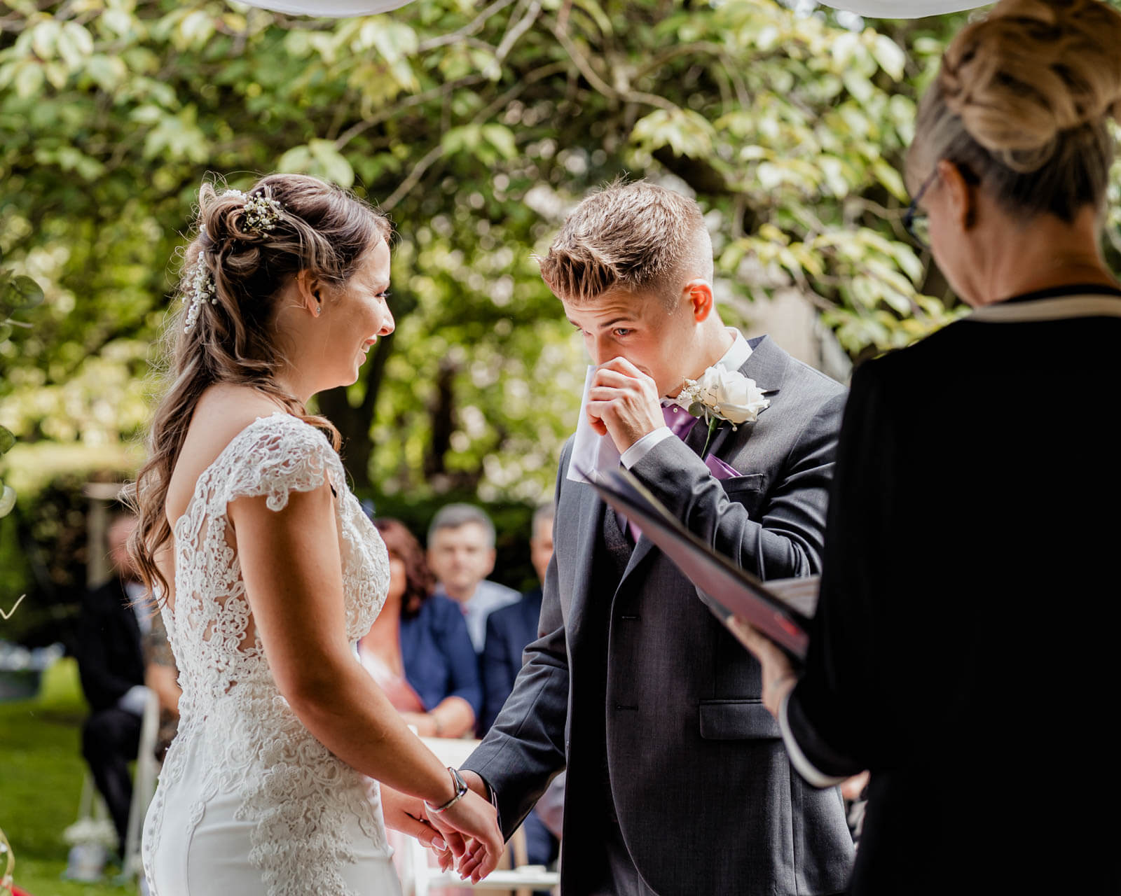 Groom wipes away tears while holding hands with bride during outdoor wedding ceremony at Hazlewood Castle