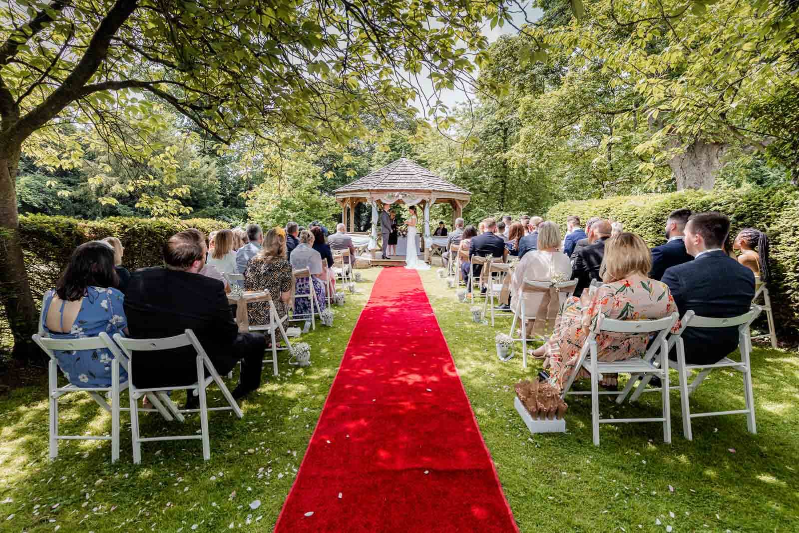 Outdoor wedding ceremony at Hazlewood Castle with guests seated along red carpet aisle leading to gazebo