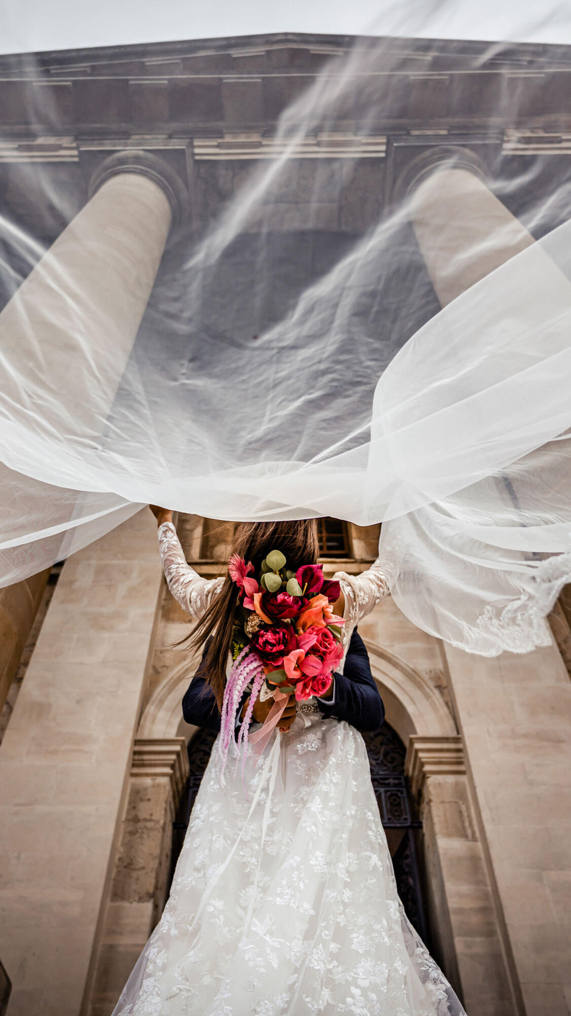 Editorial bridal portrait with flowing veil and bouquet in front of classic architecture