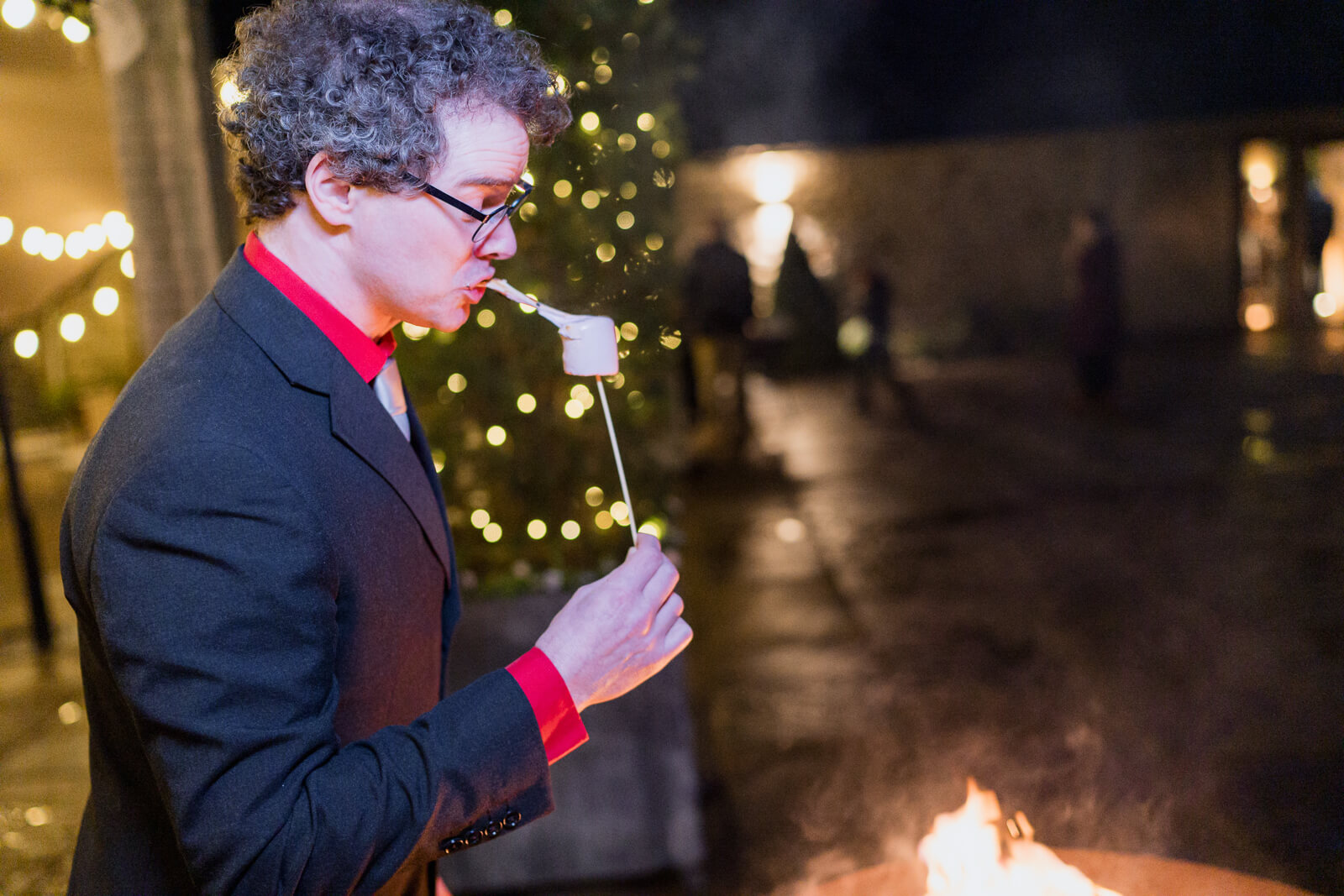 Wedding guest toasting a marshmallow over a fire pit during an evening celebration with warm lights in the background
