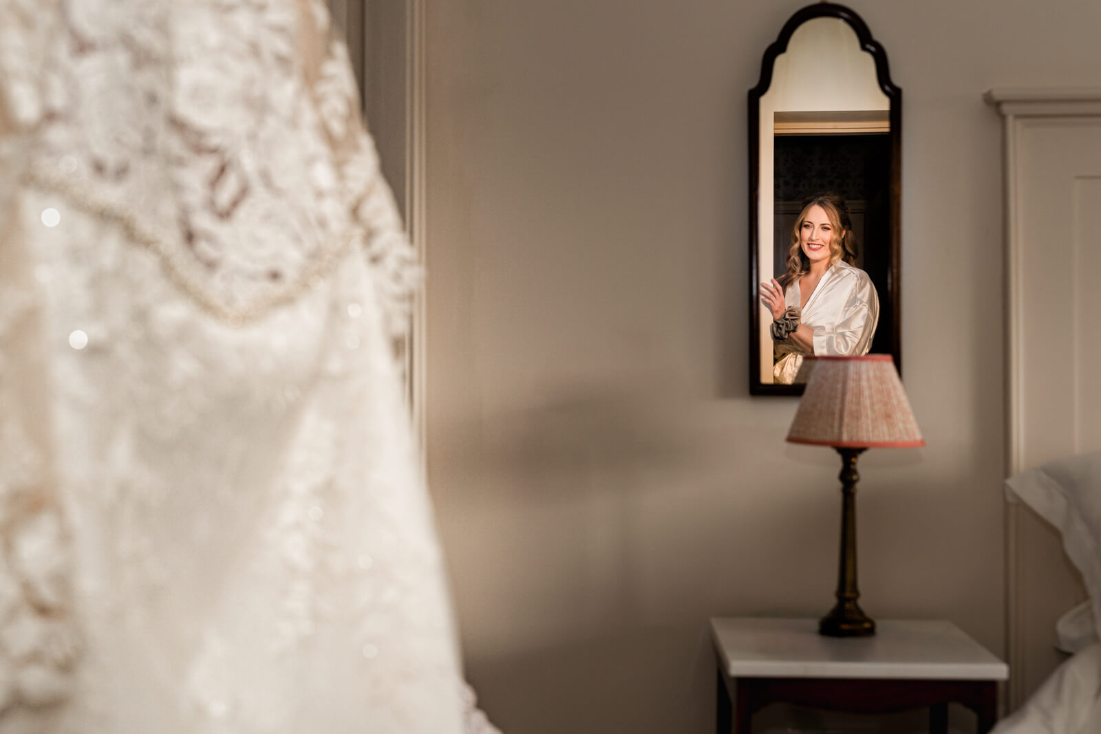 Bride smiling in mirror beside wedding dress during morning preparations at The Tithe Barn at Bolton Abbey