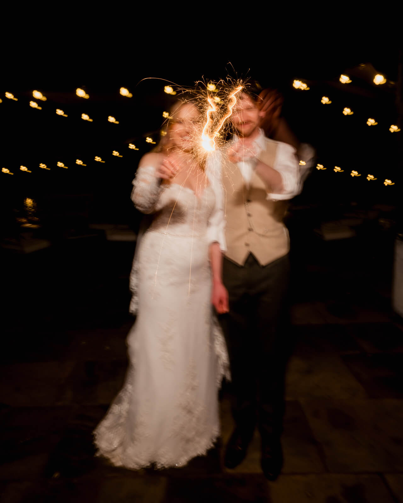 Bride and groom holding sparklers during a nighttime wedding exit with glowing lights in the background