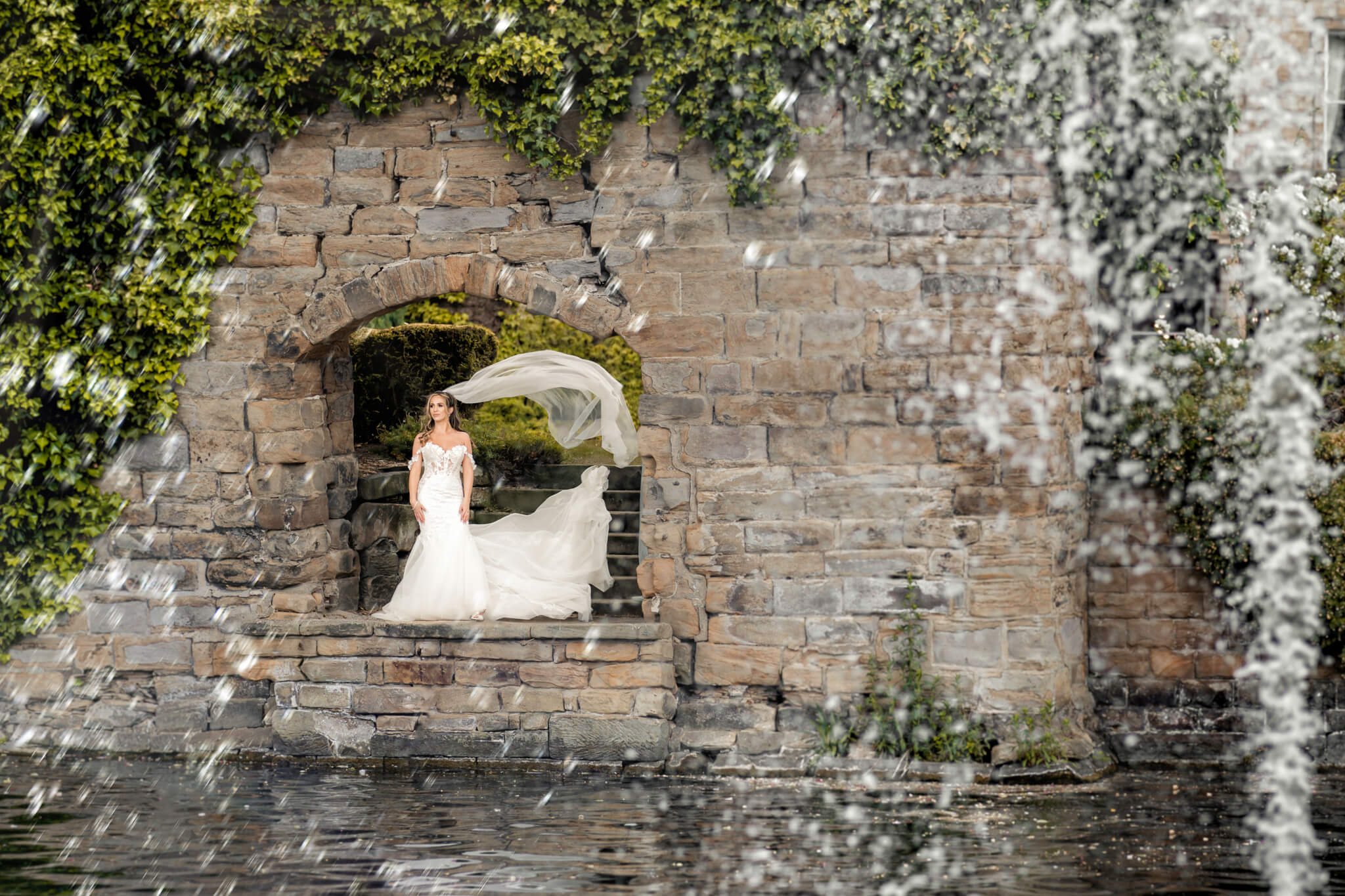 Bride standing under stone arch with veil flowing by water