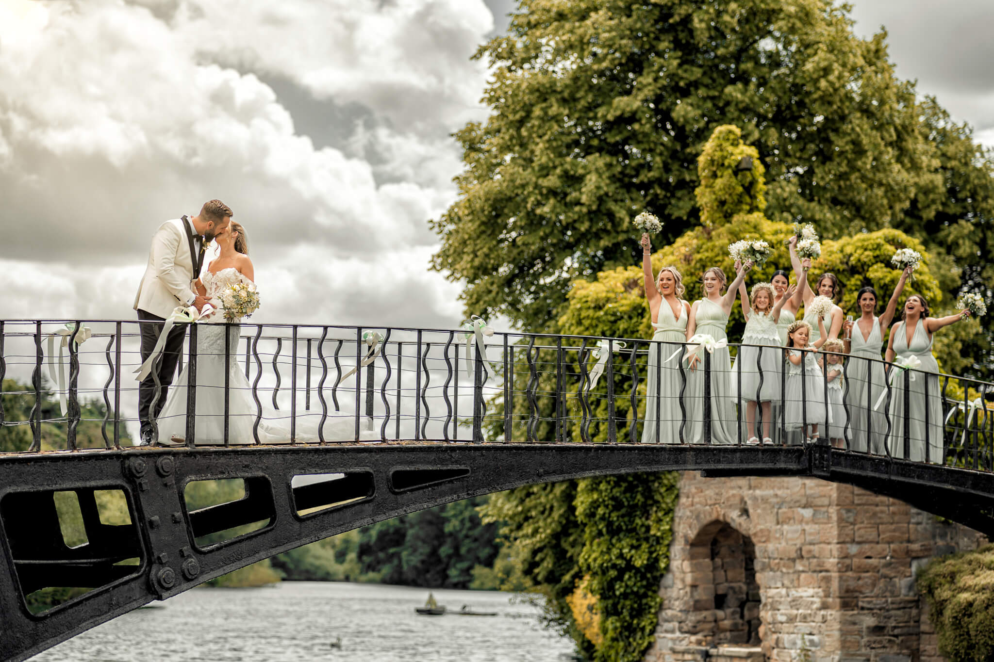 Bride and groom kissing on bridge with bridesmaids cheering nearby