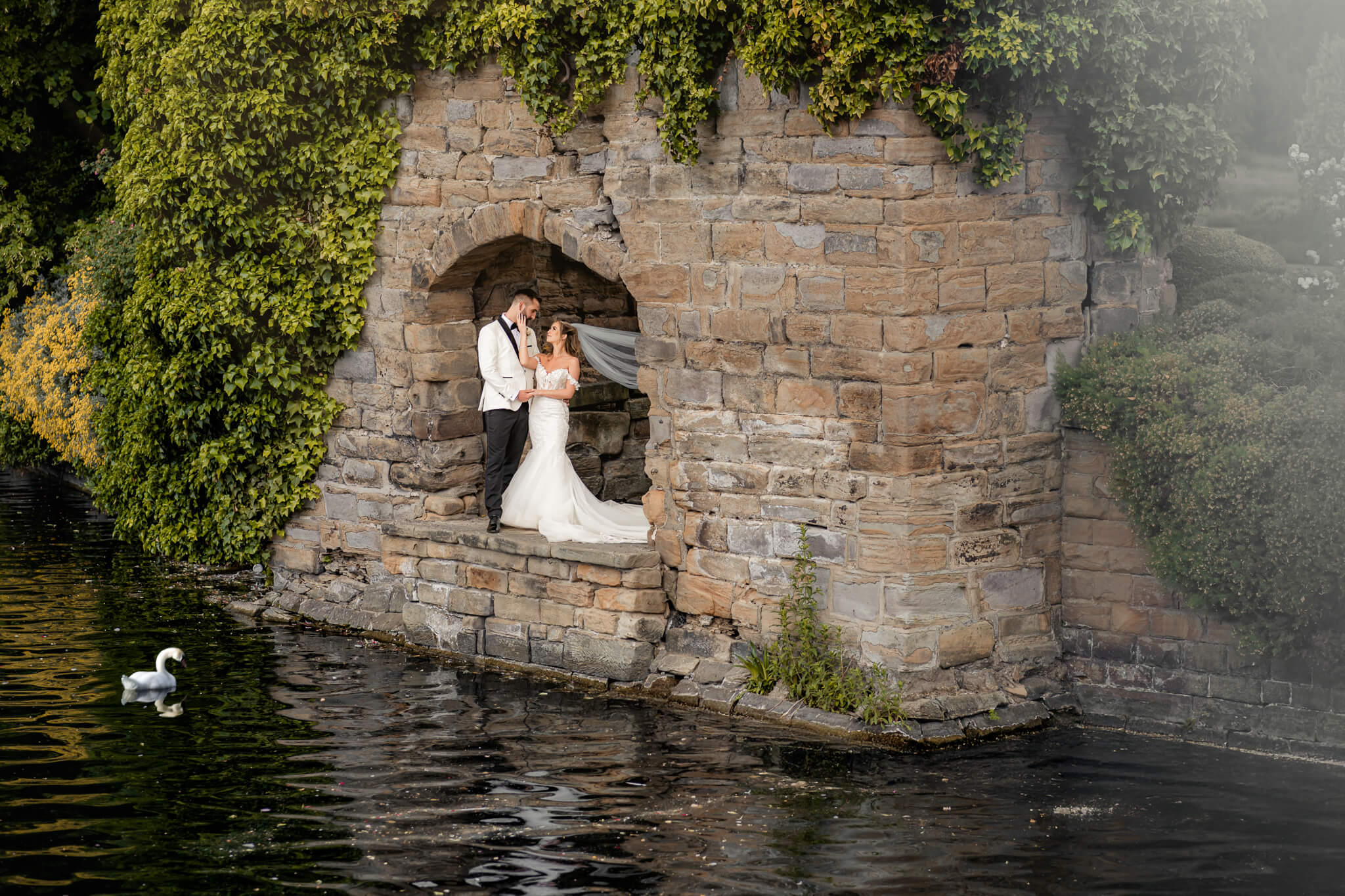 Bride and groom standing together under stone arch by lake