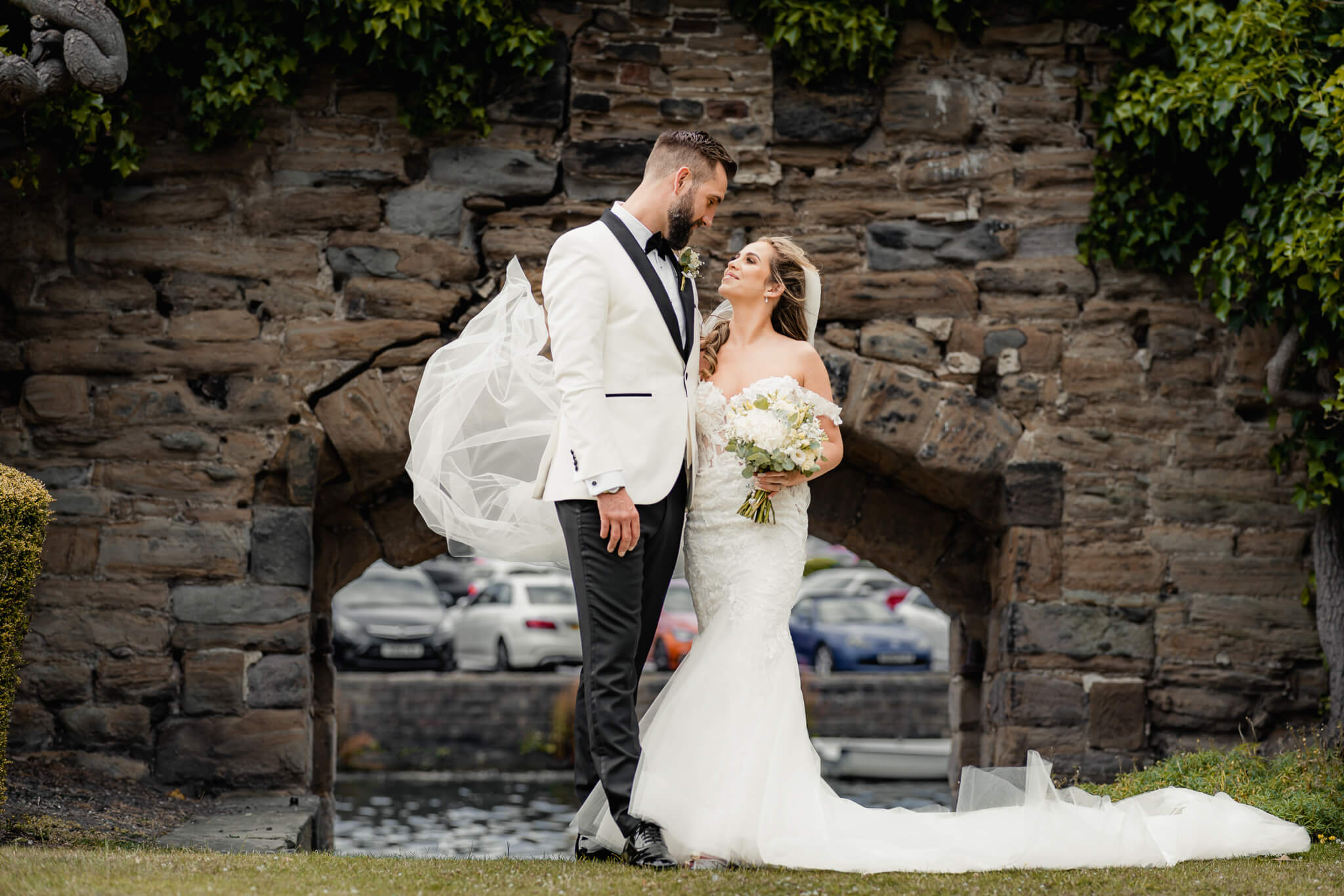 Bride and groom standing under stone arch with flowing veil