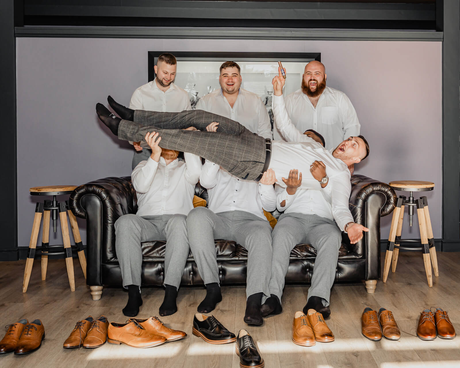 Groom being playfully lifted by groomsmen on a sofa during wedding morning preparations