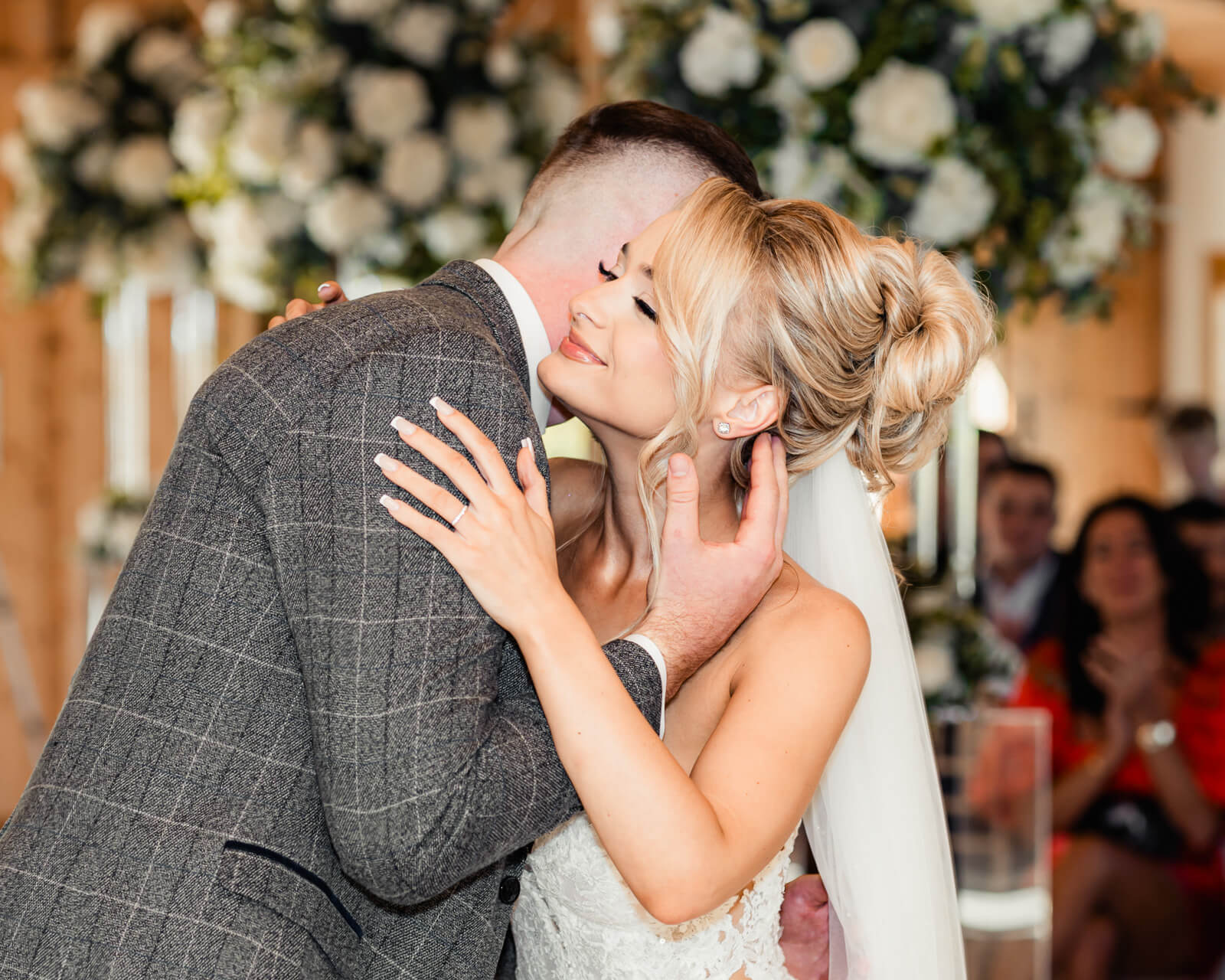 Bride smiling while embracing groom during wedding ceremony with soft floral background