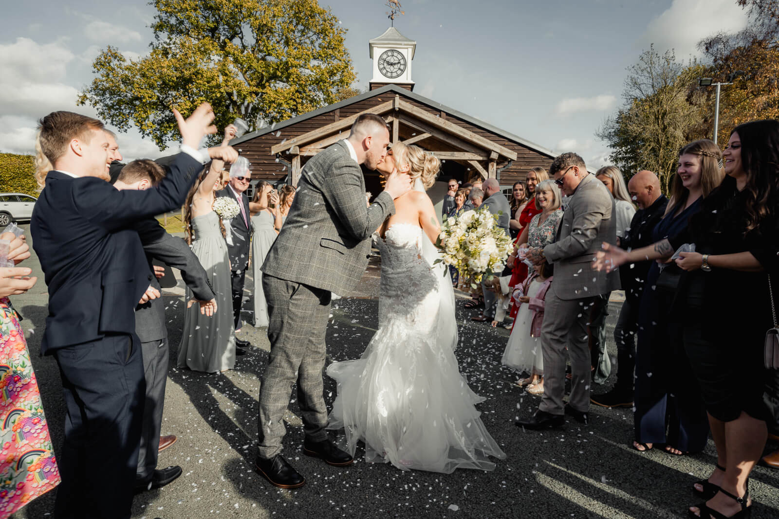 A joyful confetti moment as the bride and groom share a kiss while guests celebrate around them outside the ceremony hall in warm afternoon light.