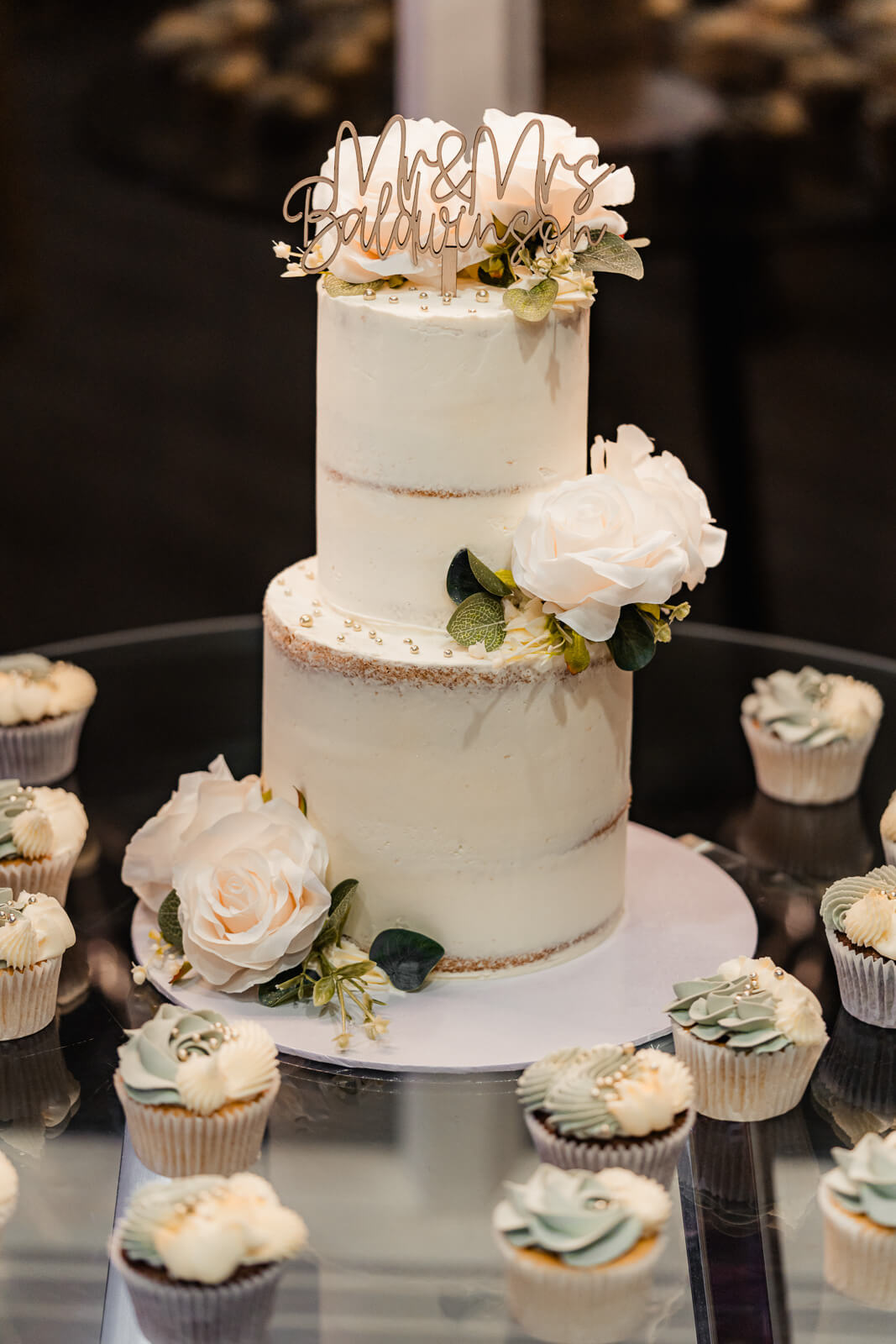 Two-tier wedding cake with white frosting, floral details, and cupcakes on display