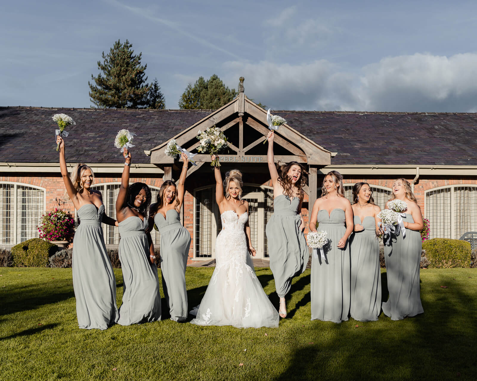 The bride and her bridesmaids celebrate together, holding their bouquets up in a joyful moment outside Colshaw Hall Country Estate.