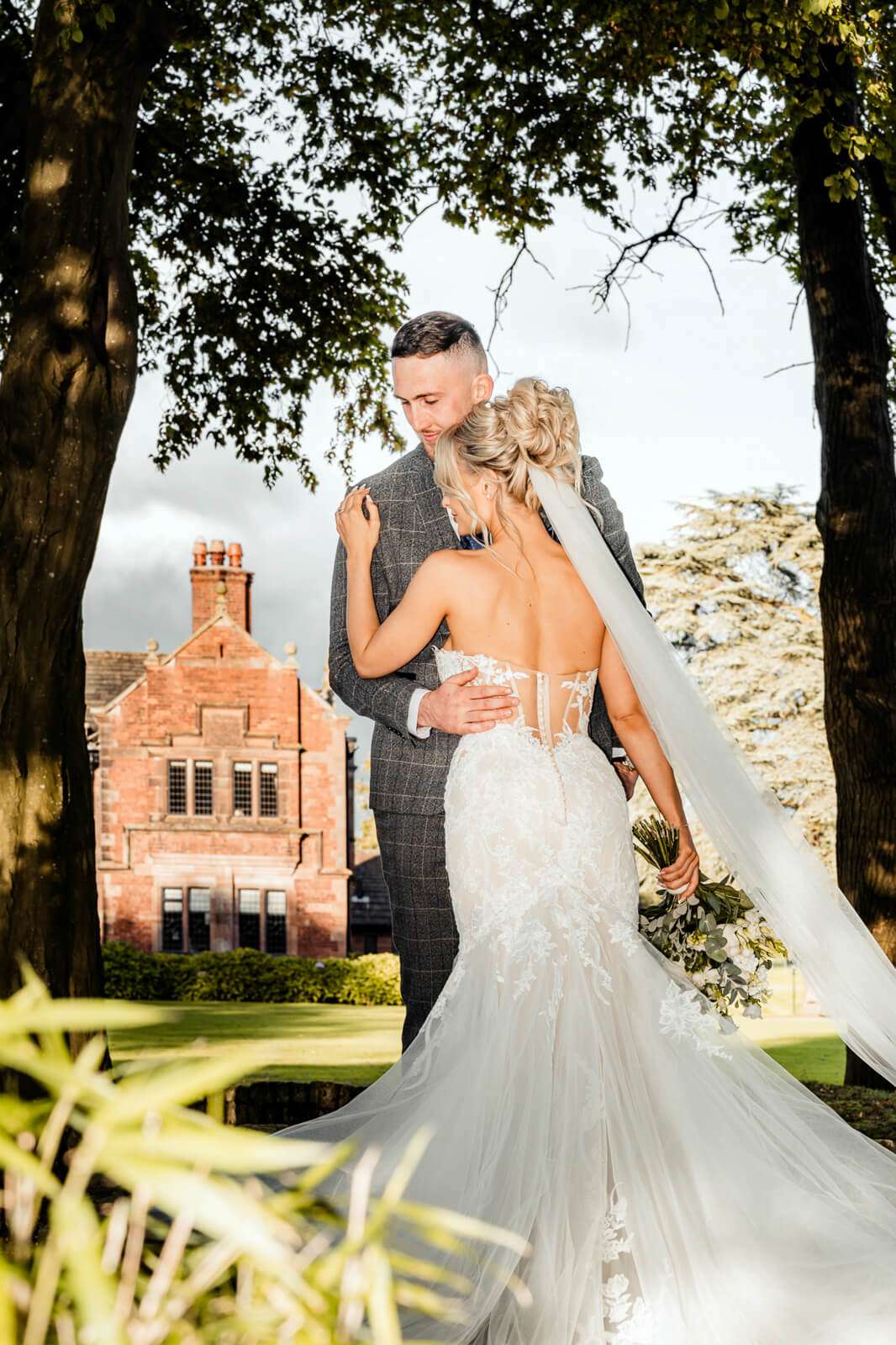 Bride and groom embracing under trees during Elegant Wedding at Colshaw Hall Country Estate with historic building backdrop
