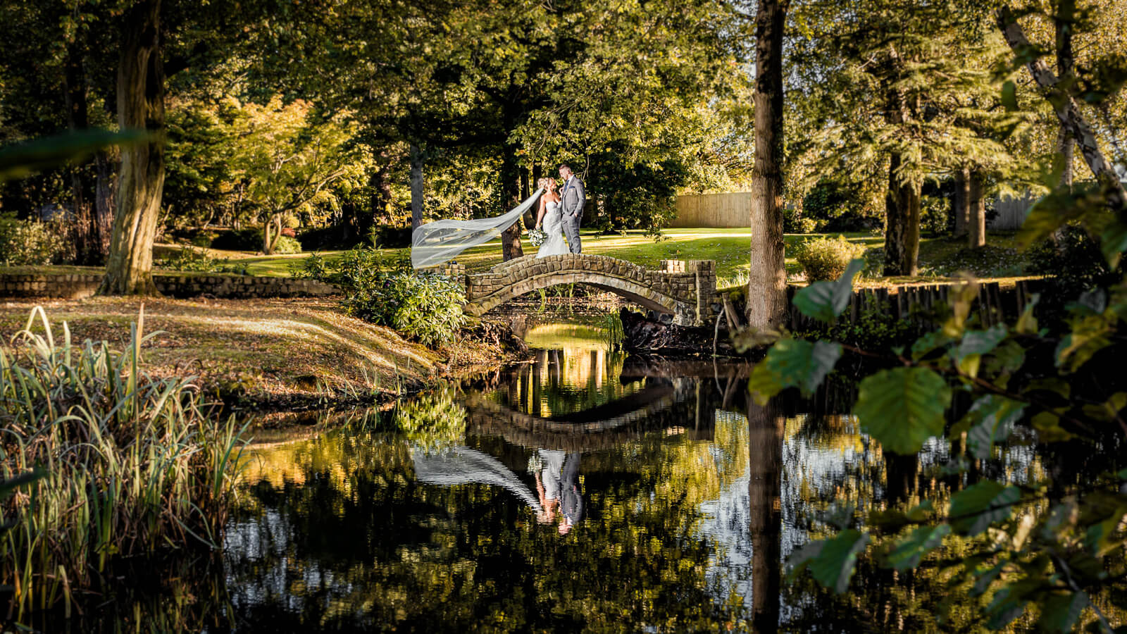 Bride and groom on stone bridge during Elegant Wedding at Colshaw Hall Country Estate with reflection in water and flowing veil