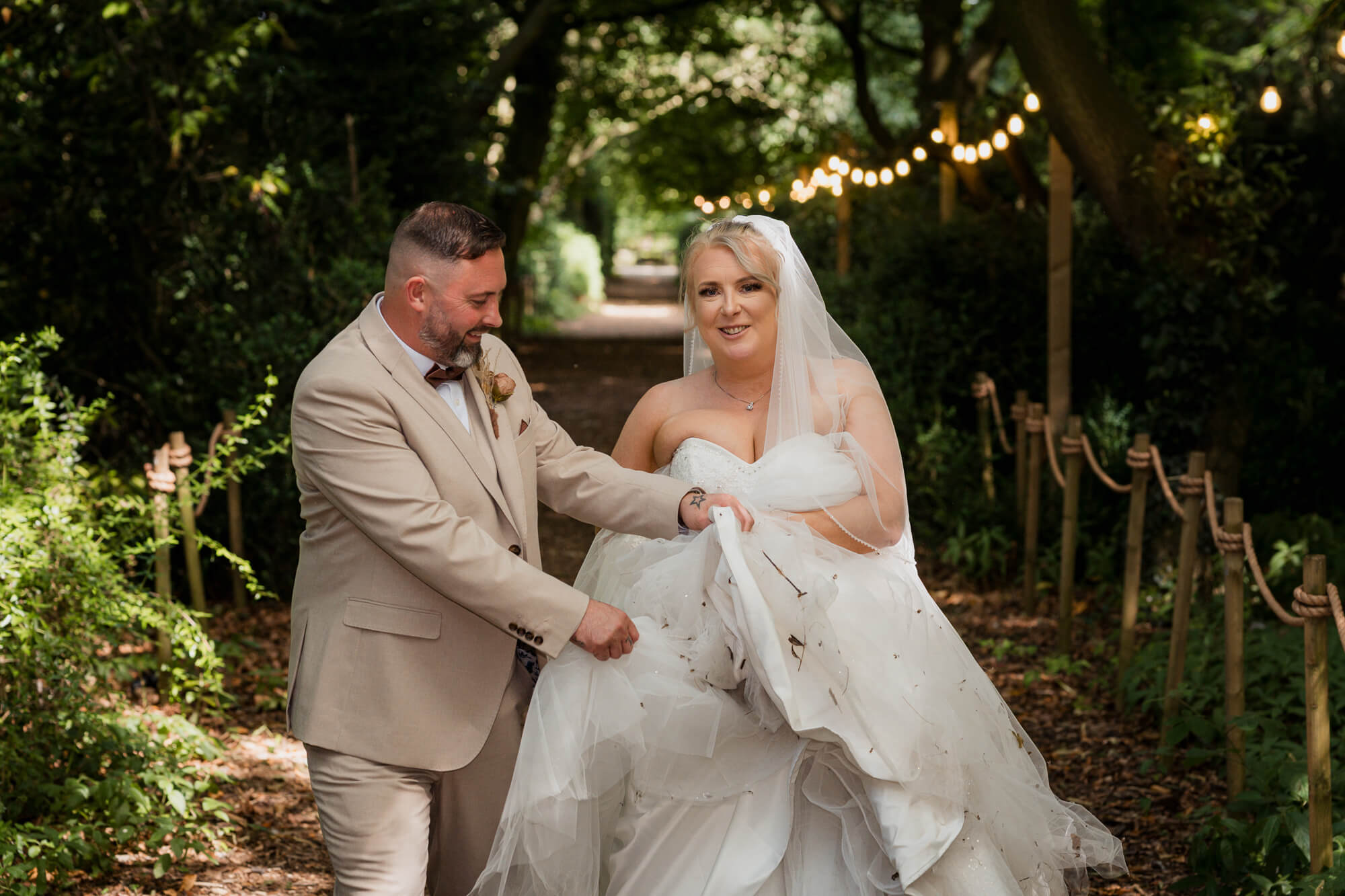 Groom helping lift the bride's wedding dress along the woodland path.