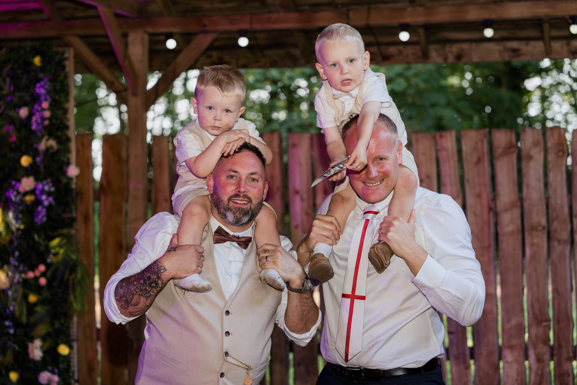 Two fathers carrying young children on their shoulders during the evening wedding celebration.