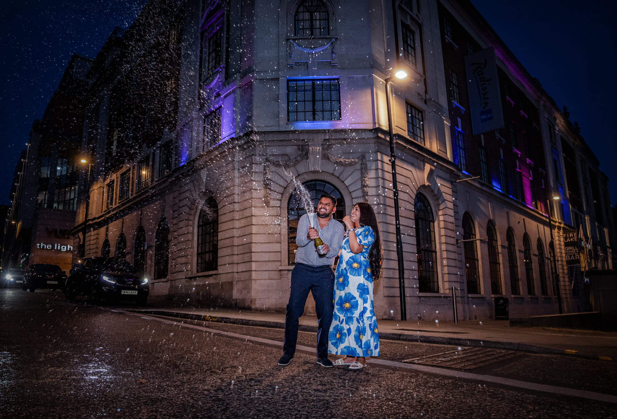 Couple celebrating with a champagne spray outside Radisson Blu Hotel during a Leeds pre-wedding photoshoot at night.