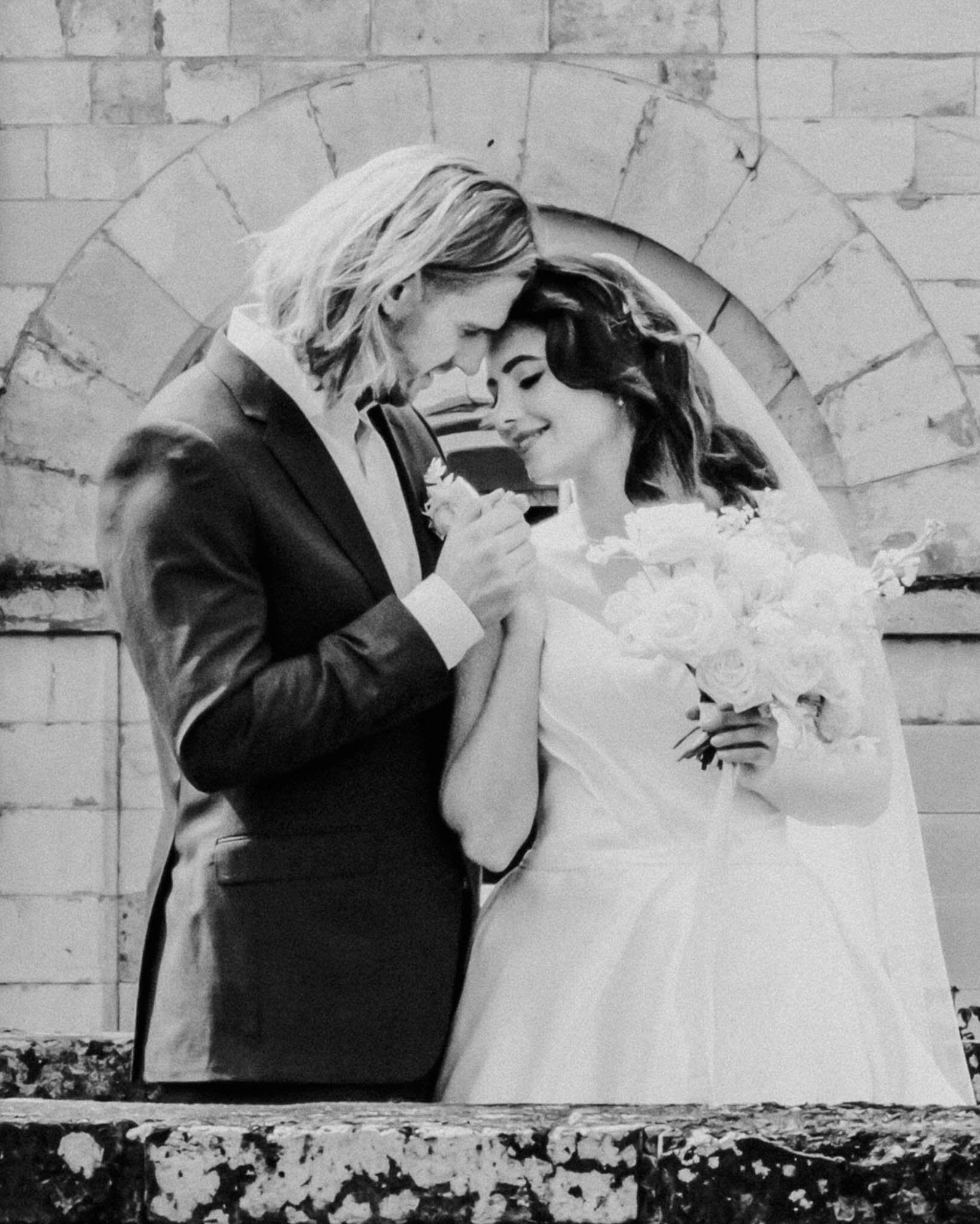 Groom facing the bride and holding her hand into his palm while she is holding the bridal bouquet close to her chest. This is a romantic moment on the bridge of Hazlewood Castle. 