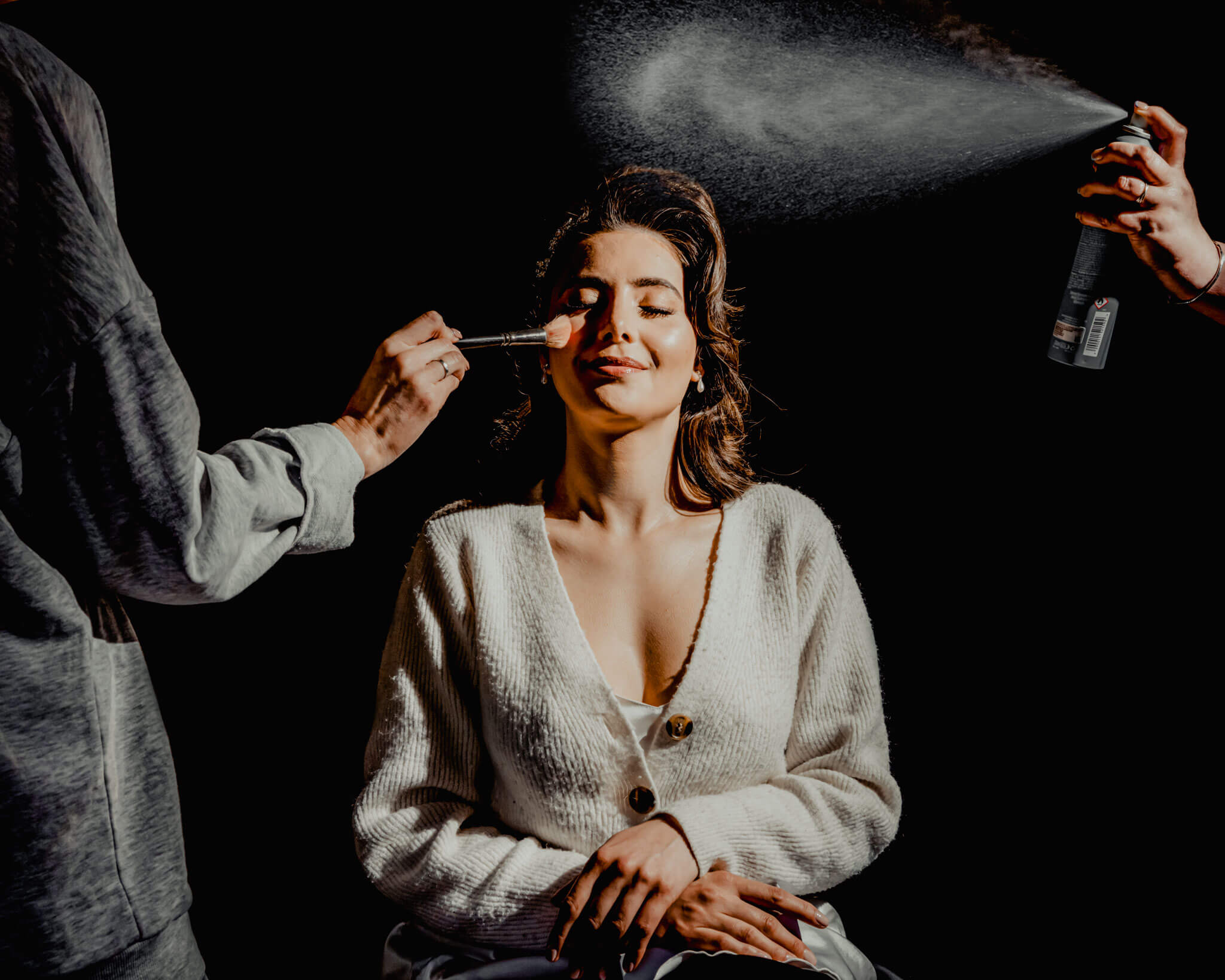 Bride getting her make-up and hair done at Hazlewood Castle. We can see her beautiful smile and only the hand with the brush of the make-up artist to the left and the hand of the hair stylist spraying her hair to the right.