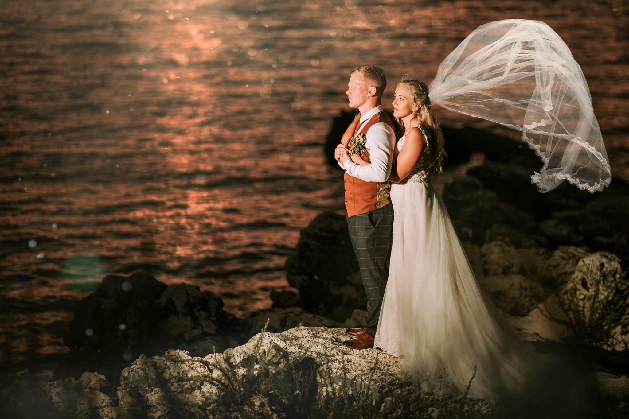 Bride holding the groom into her arms from behind looking towards the sea.